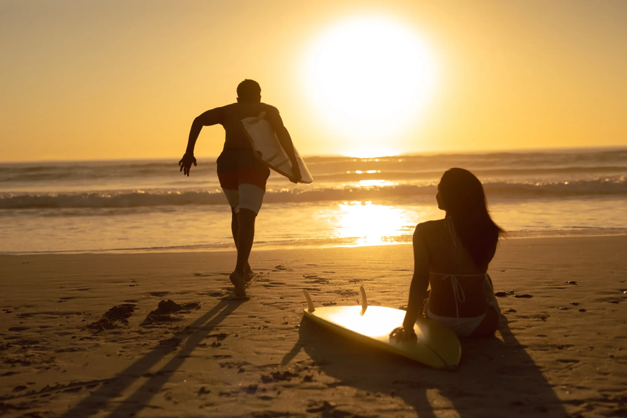 Hombre corriendo con tabla de surf mientras mujer se relaja en la playa. Ilustra como reducir el estrés y la ansiedad con hábitos saludables